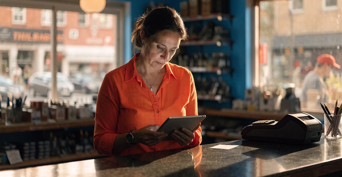 Small business owner in an orange shirt reviewing a tablet behind the counter of a hardware store with a cerulean blue wall.